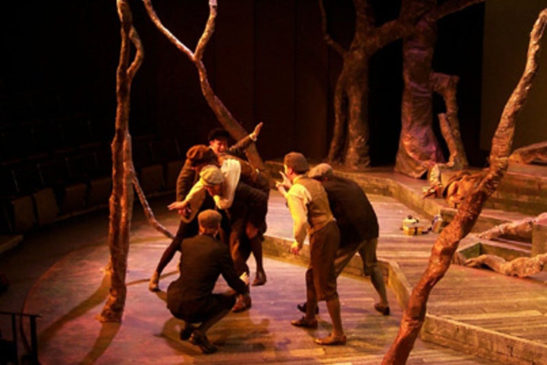A person carrying another on their back while surrounded by four men on a forest-themed stage
