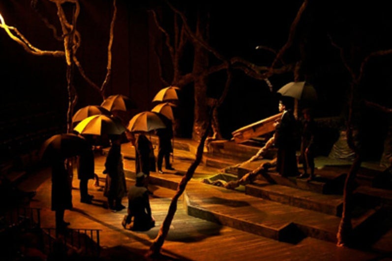 Group of people on forest-themed stage with umbrellas looking at a man at the top of stairs