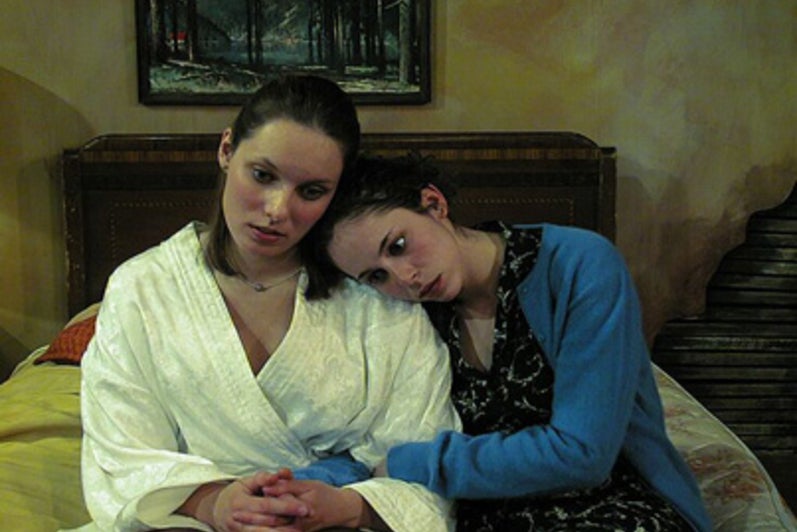 Two women sitting on a bed with one's head on the other's shoulder
