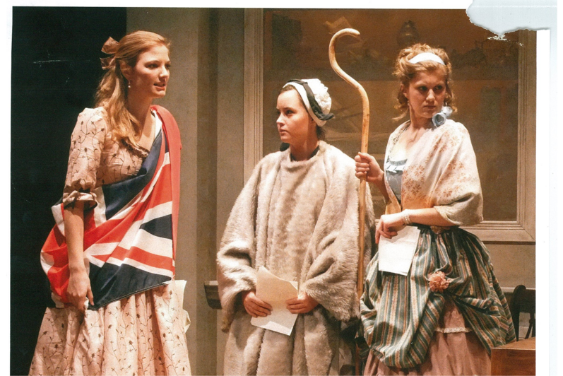 One woman in a large sash with the British flag, a woman with white shawl and a woman holding a wooden staff