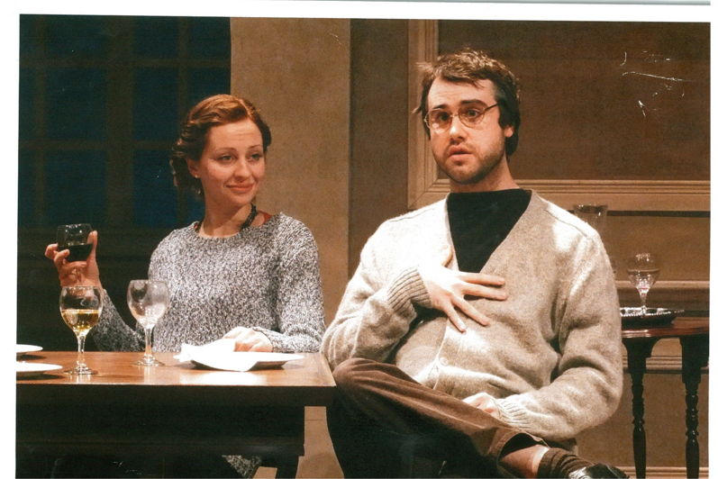 A woman sitting with a glass wine and a man speaking to someone off-screen