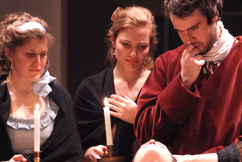 Two women holding candles looking remorseful and a man examining body