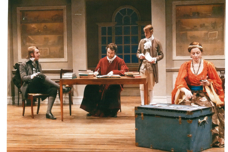 Man in red cloak signing documents while conversing with two men, and a woman sits infront holding a glass of wine
