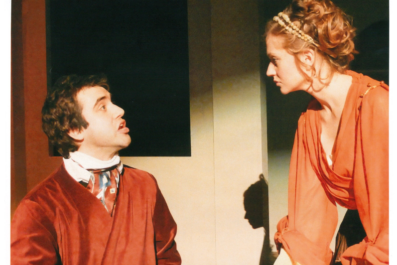 A man in red cloak (sitting) speaks to woman in orange blouse (standing)