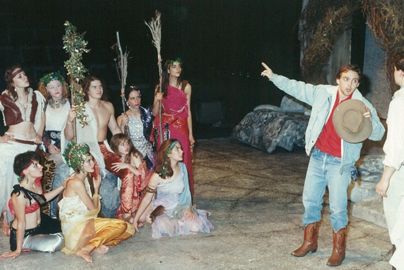 Man dressed in modern clothes gesturing to a group of nine people in colourful clothing