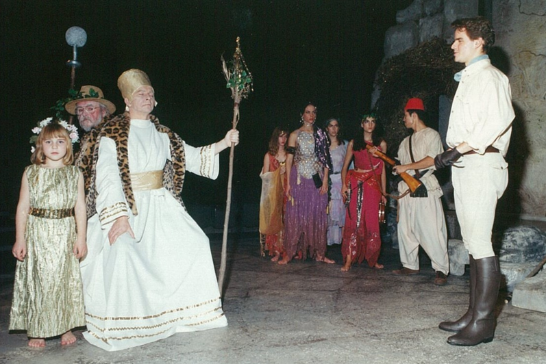 Man with a crown and large staff with two people standing behind him, a group of colourfully-dressed people and a man in an European explorer outfit