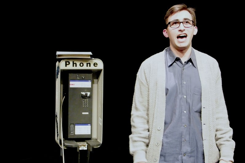 Simon stands on stage beside a pay phone