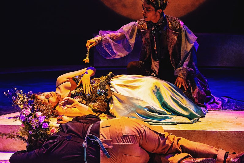 Oberon (Linus Ma, top) holds a flower over a sleeping Titania (Morgaine McEvoy, middle) to undo her spellbound love to the donkey-headed Nick Bottom (Theo Patterson, below). The scene is bathed in golden light with a large orange moon in the background.