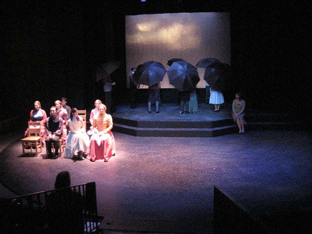 Group of people sitting on stage with another group in the background holding umbrellas