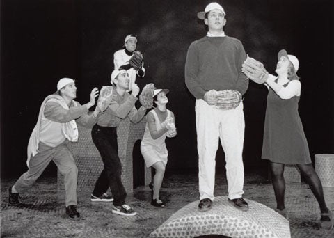 Man in foreground with a woman standing to his right, and four others looking at him in the background while they all wear baseb
