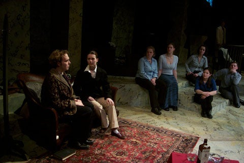 Two people sitting and talking to each other with the rest of the cast looking on in the background