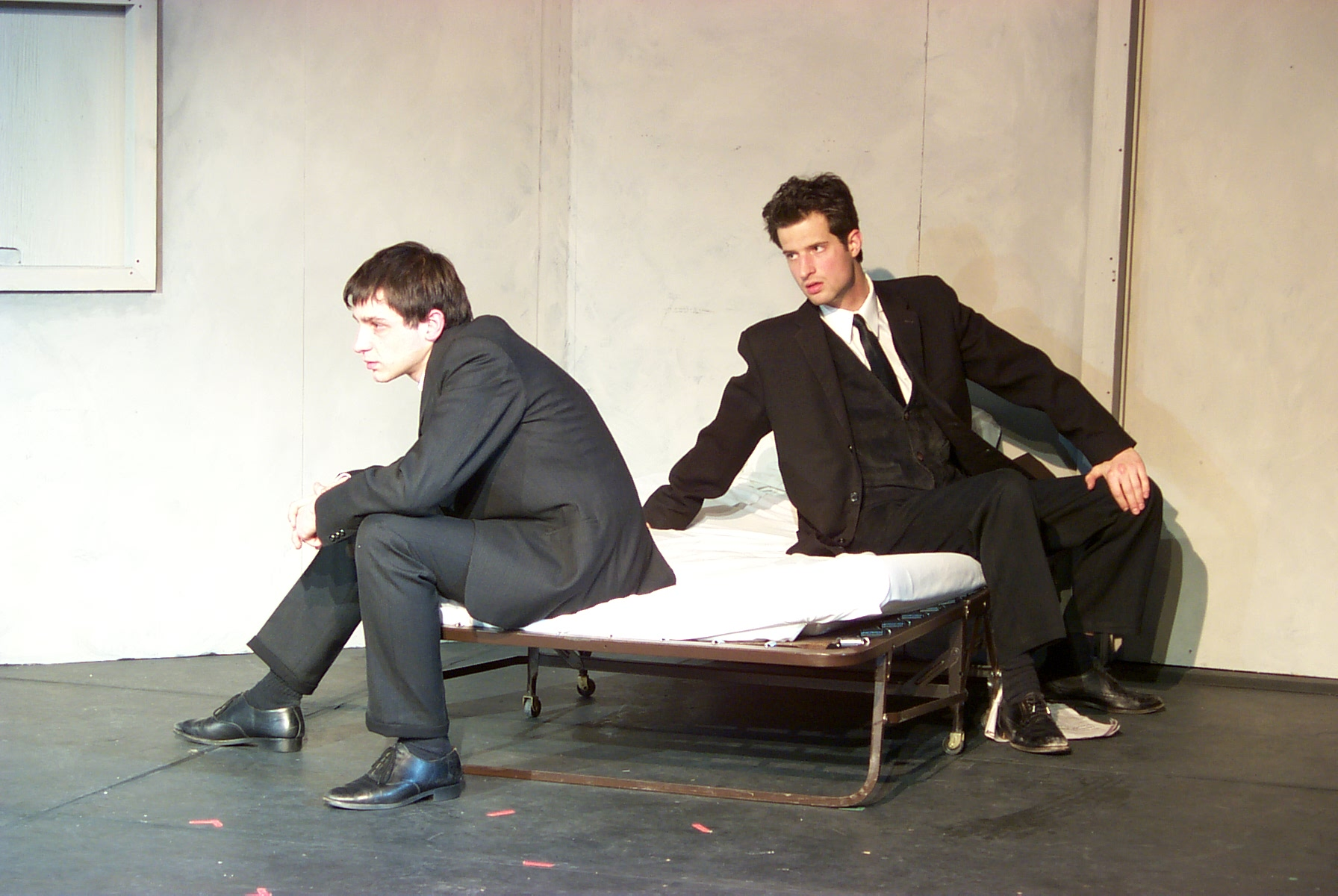 Two men in black suits sitting on a bed, man on the right looks to the one on the left