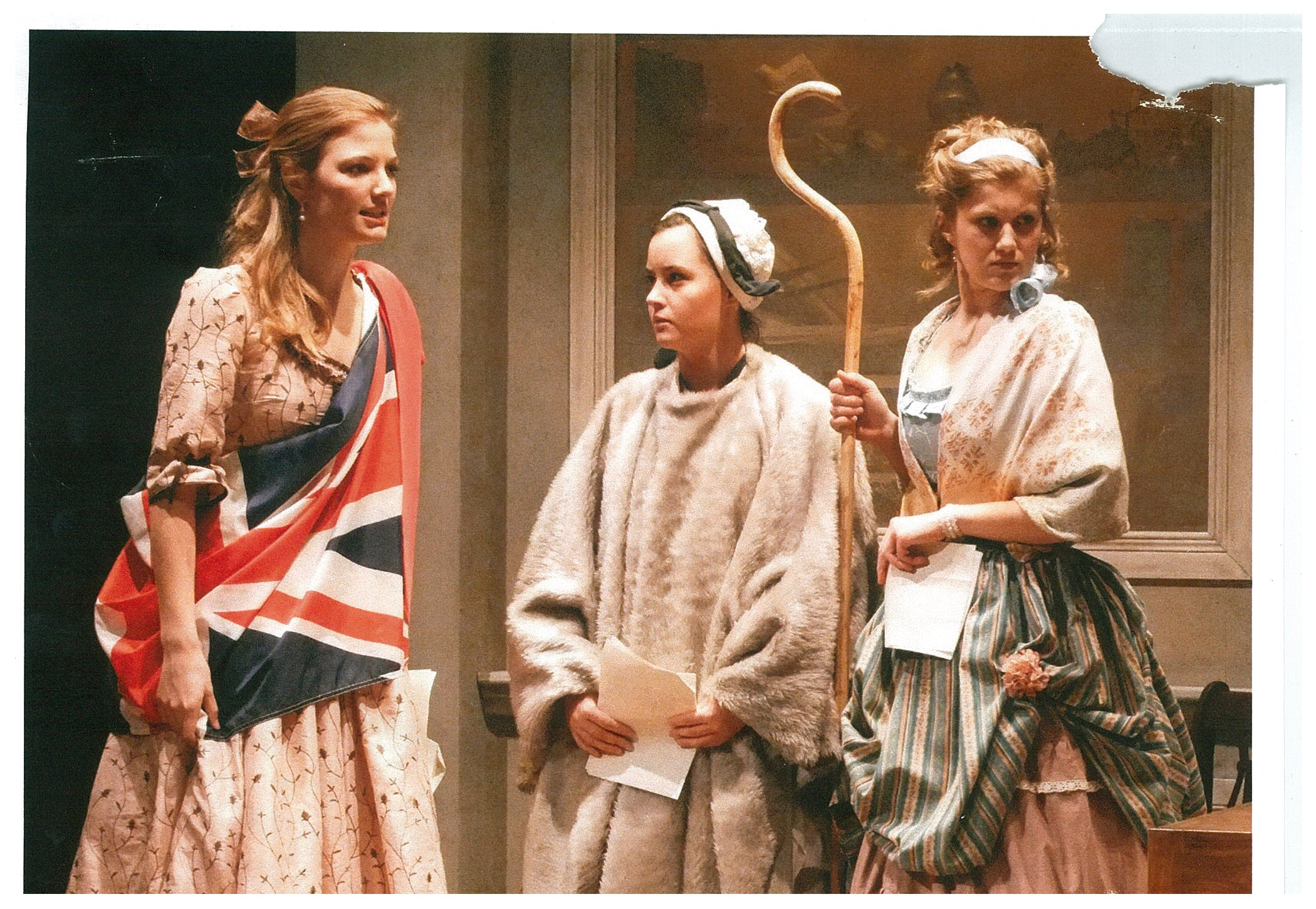 One woman in a large sash with the British flag, a woman with white shawl and a woman holding a wooden staff
