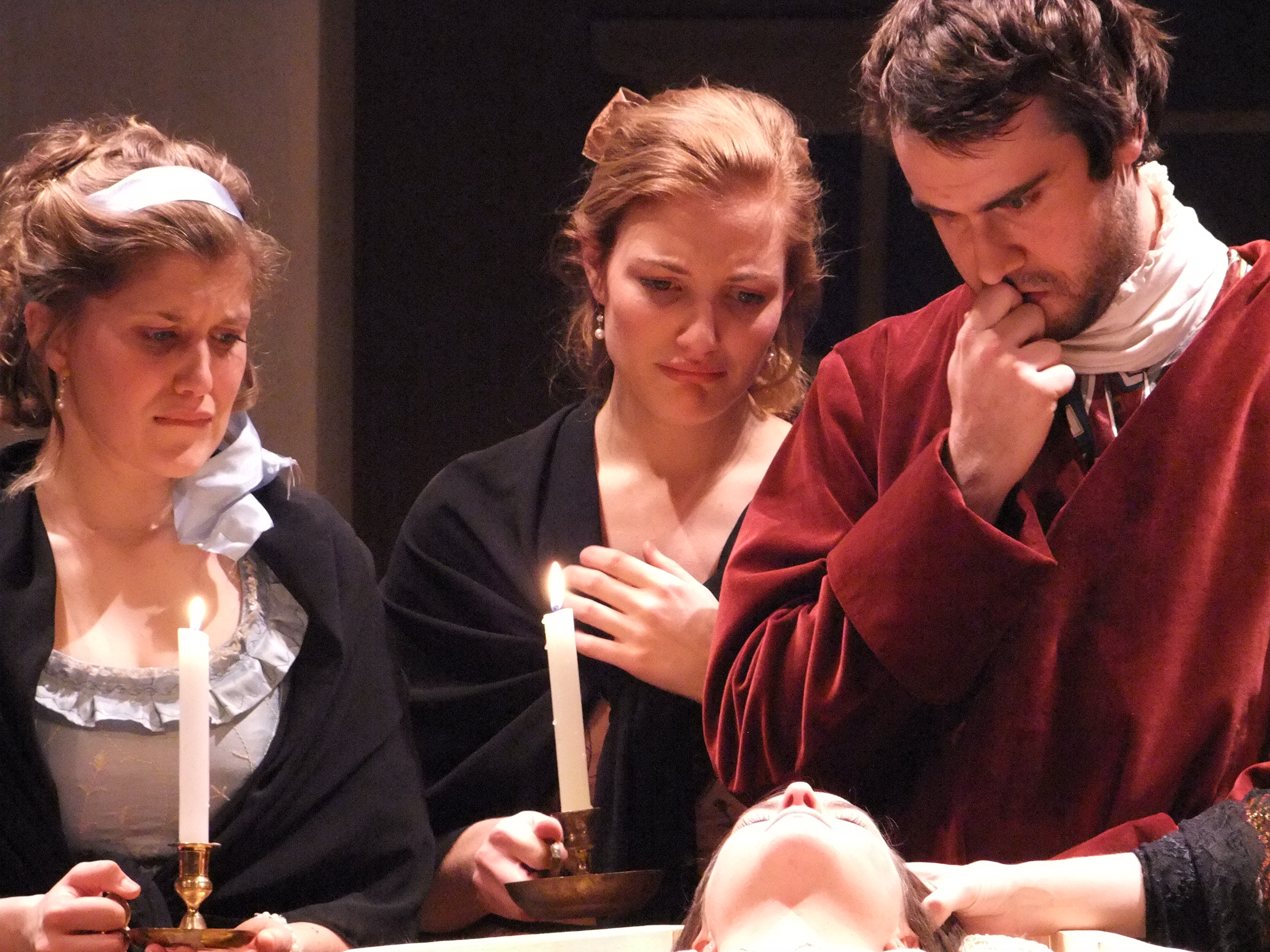 Two women holding candles looking remorseful and a man examining body