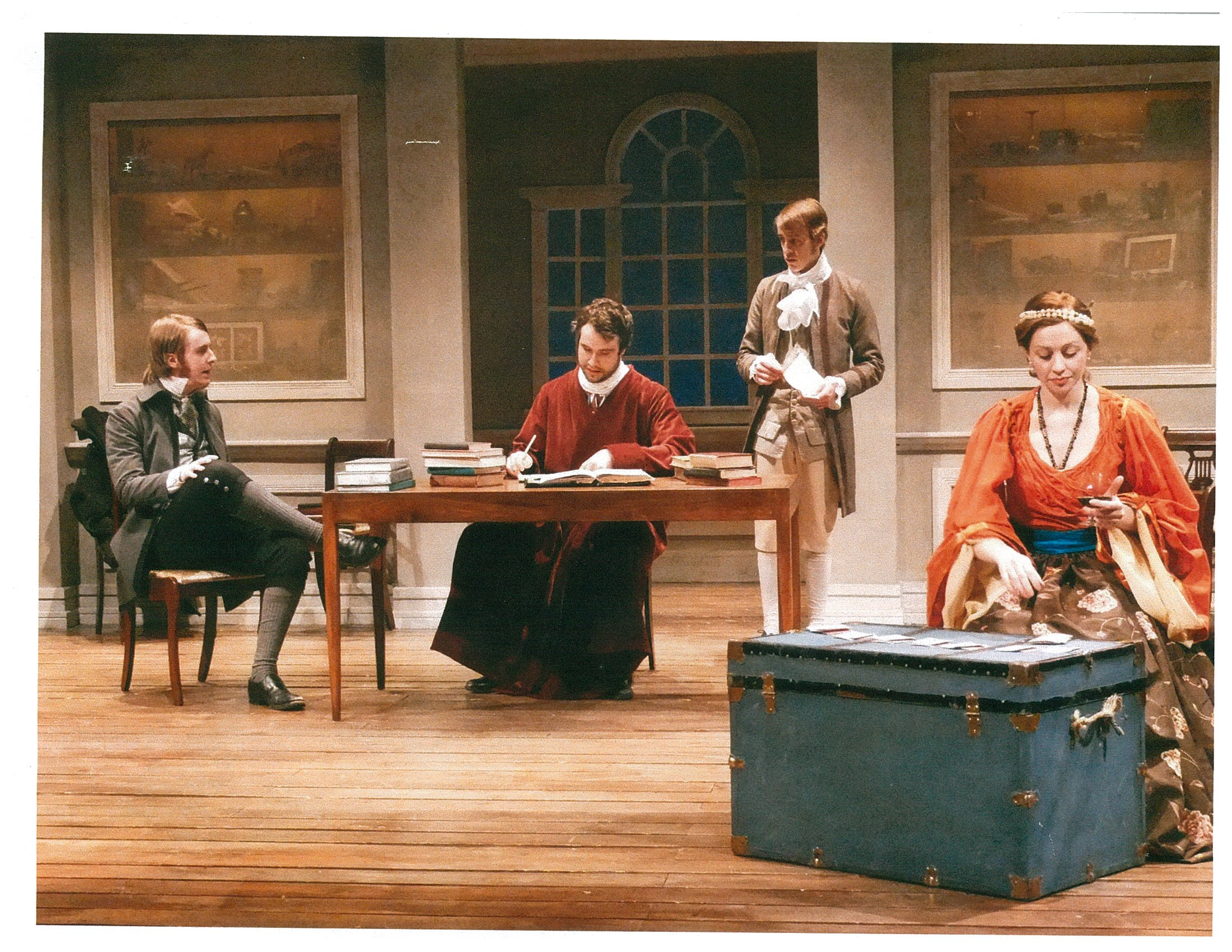 Man in red cloak signing documents while conversing with two men, and a woman sits infront holding a glass of wine