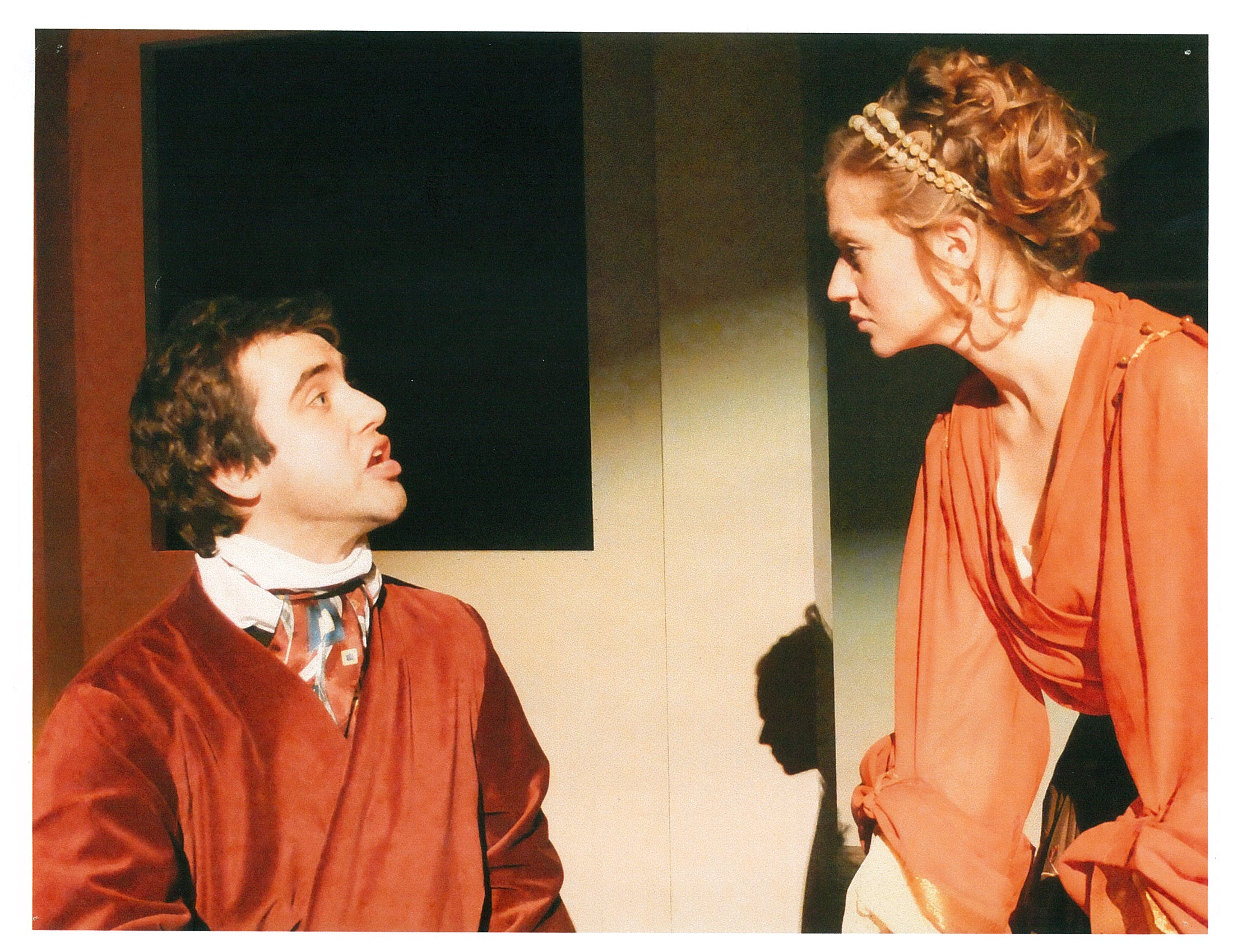 A man in red cloak (sitting) speaks to woman in orange blouse (standing)