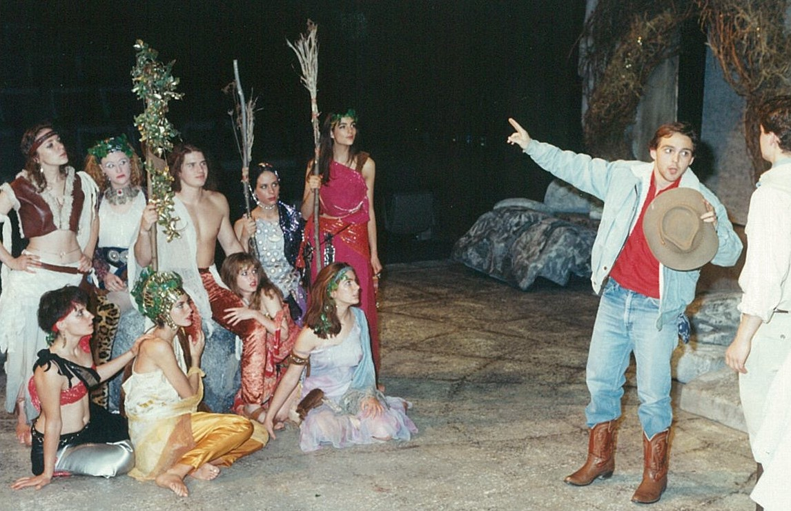 Man dressed in modern clothes gesturing to a group of nine people in colourful clothing