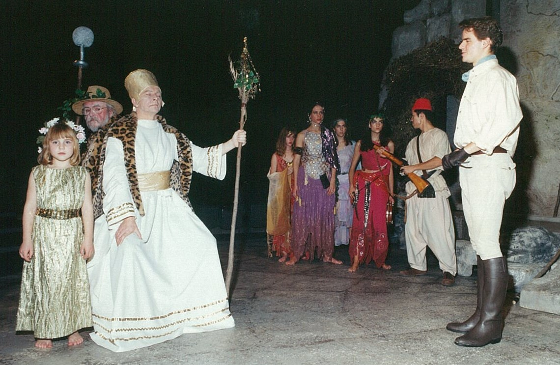 Man with a crown and large staff with two people standing behind him, a group of colourfully-dressed people and a man in an European explorer outfit