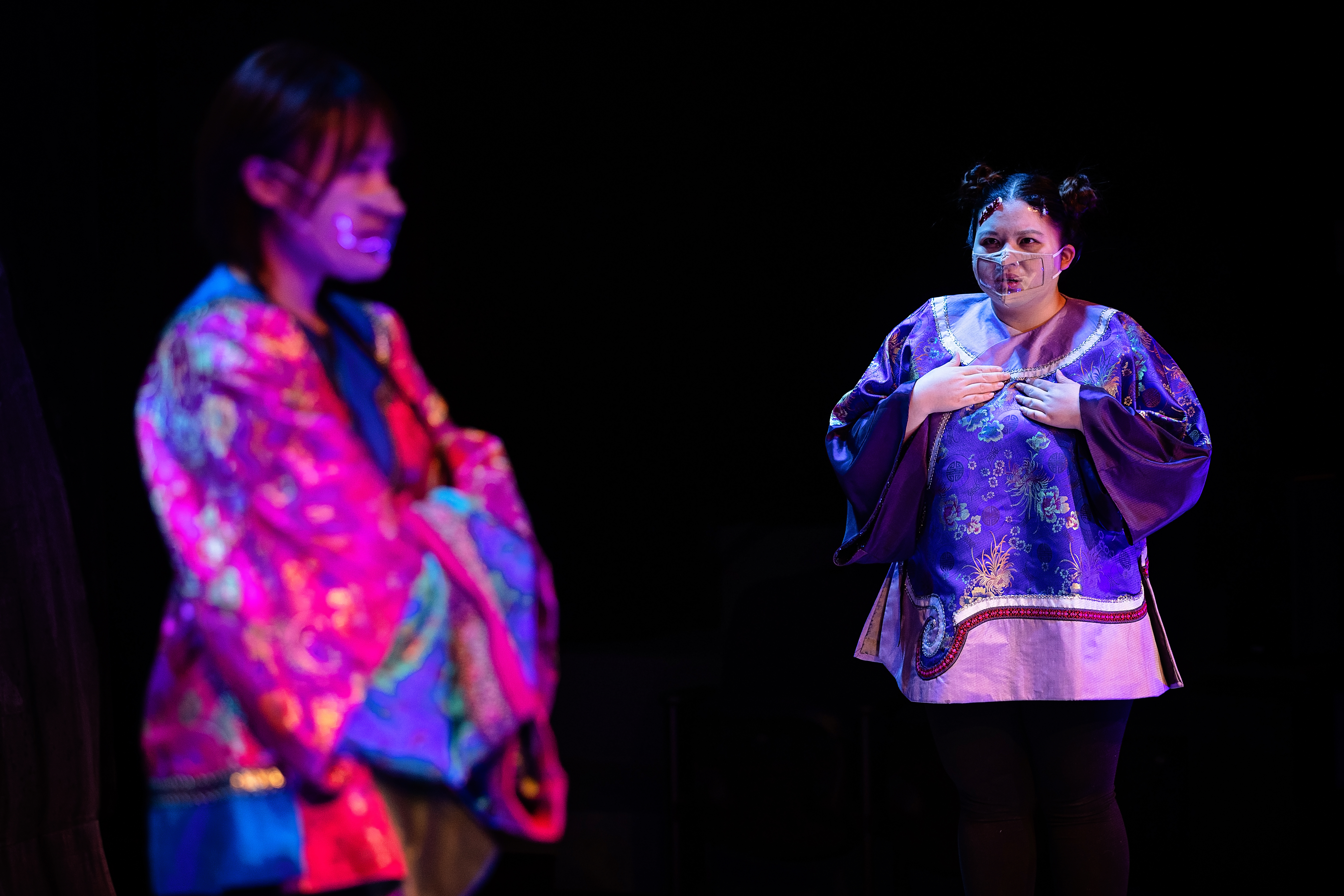 Two women wearing masks, one woman holds her hands to her chest, staring at the second woman