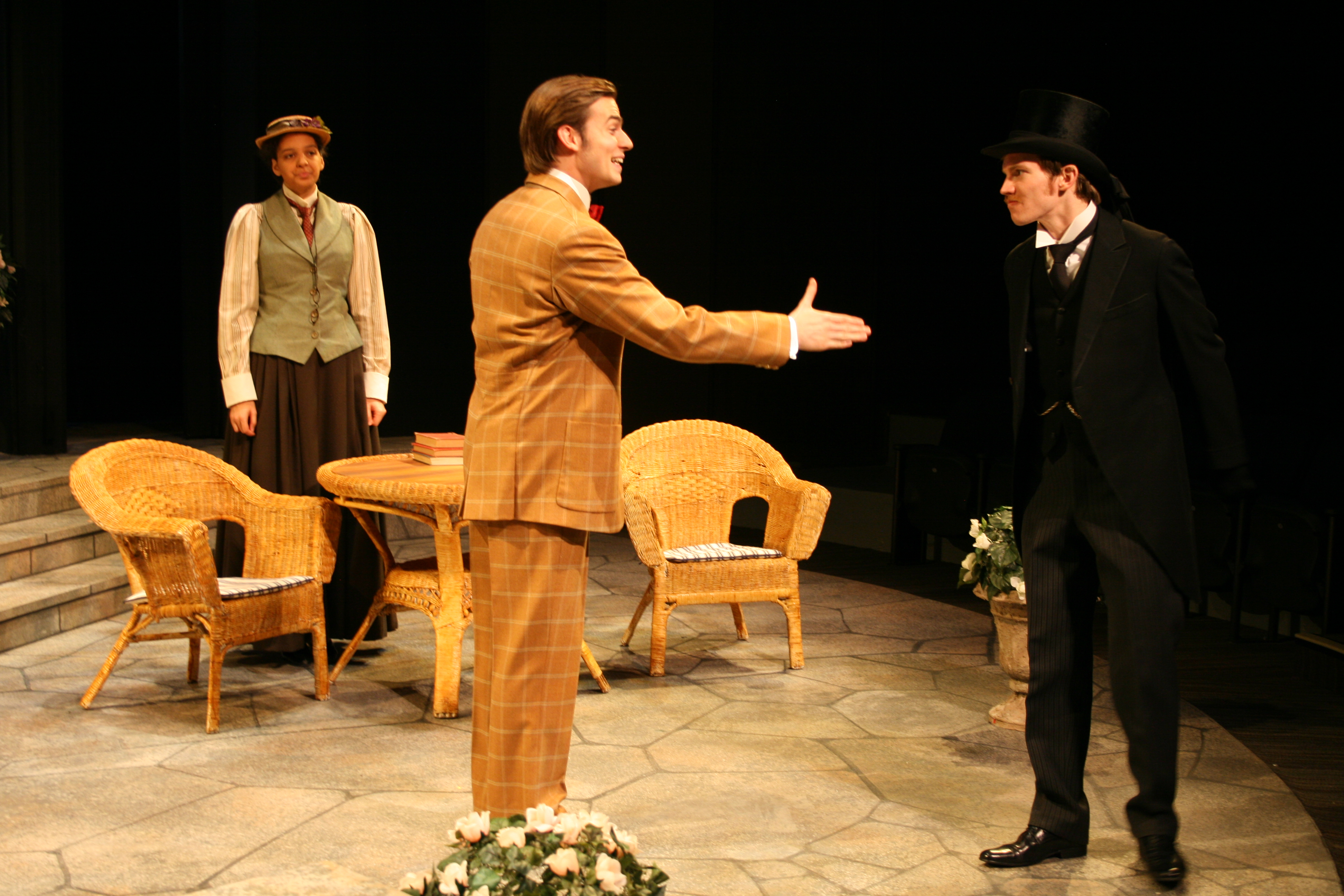 Man in brown suit holds up hand to second man in black suit, while a woman watches in the background