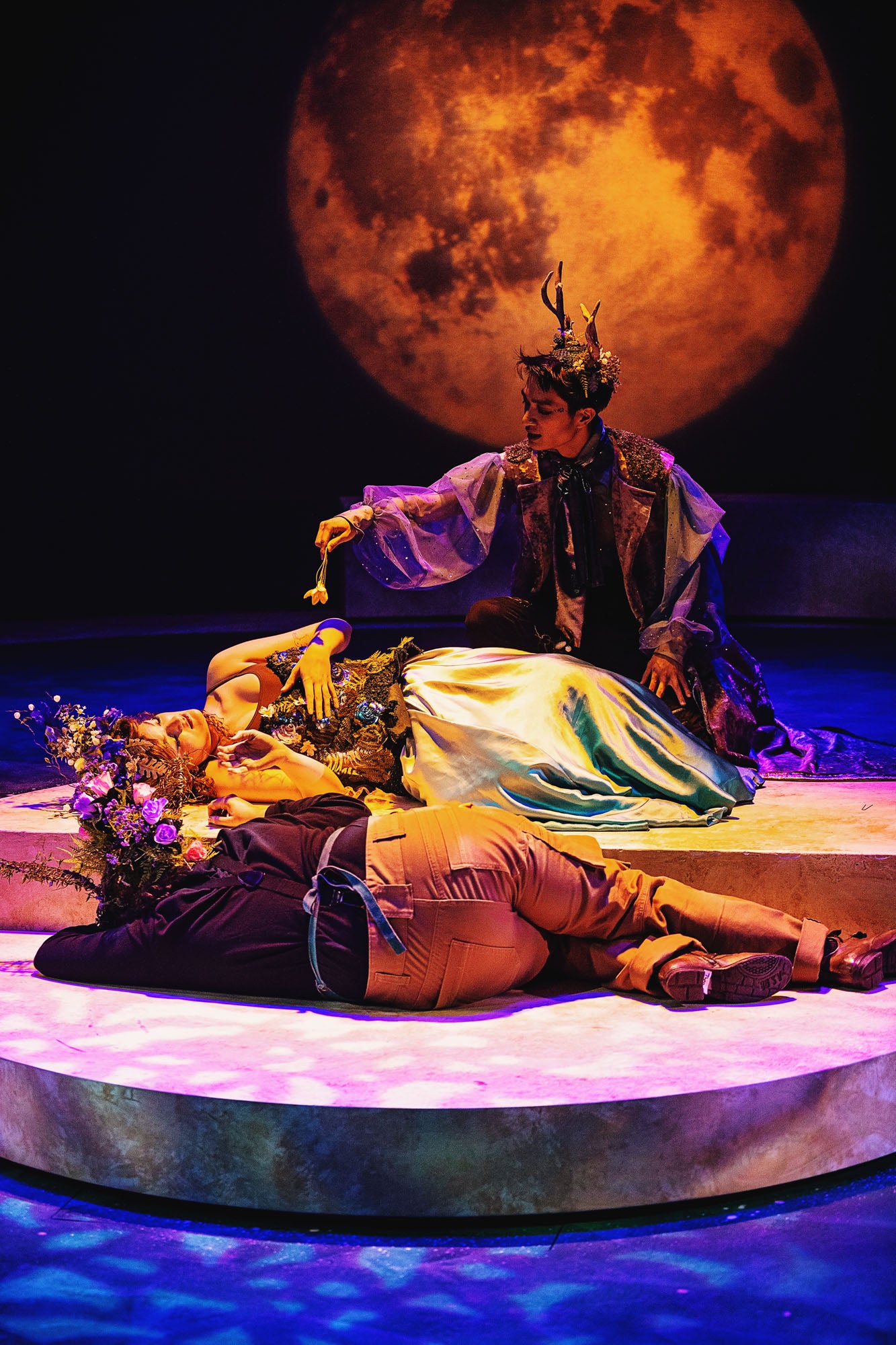 Oberon (Linus Ma, top) holds a flower over a sleeping Titania (Morgaine McEvoy, middle) to undo her spellbound love to the donkey-headed Nick Bottom (Theo Patterson, below). The scene is bathed in golden light with a large orange moon in the background.