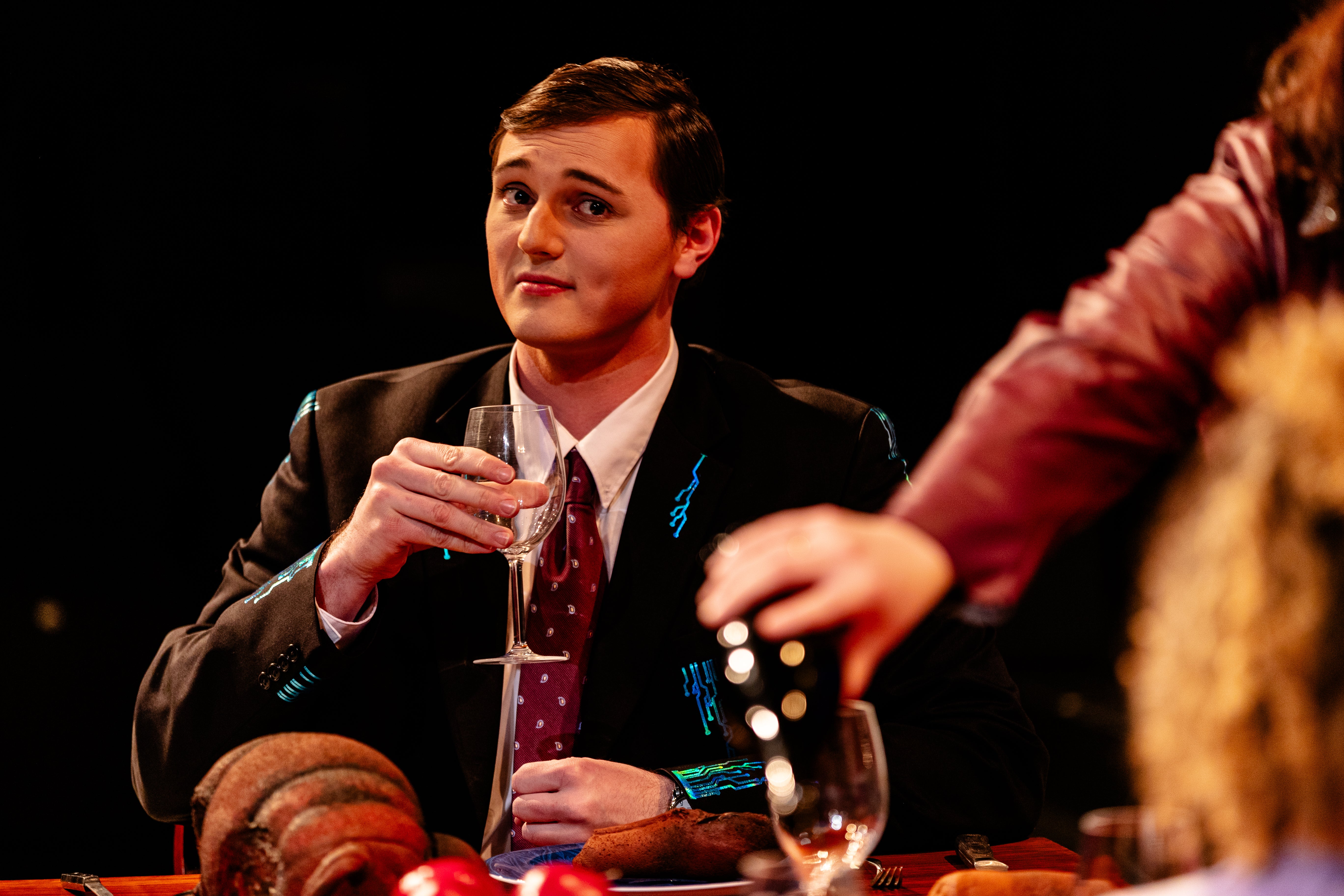 Johnny holding a glass of wine, sitting at dinner table with other characters