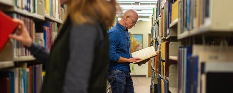 An older man, an MTS grad student, reads a book in the library stacks at Grebel. A woman pulls a book out of a shelf closer to the camera.