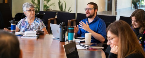 A student speaks during a class, sitting around a large round table with class mates.