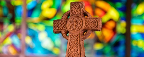 A stone celtic cross stands in front of a colourful stainglass background