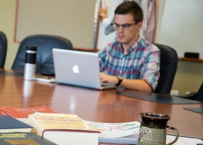 Student typing on laptop with theology books on table