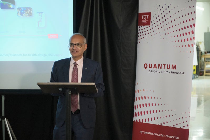 A person standing in front of a red and white banner and a projector screen, giving a talk. 