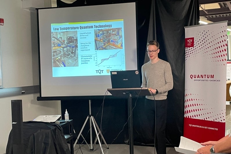 A person standing in front of a red and white banner and a projector screen, giving a talk. 