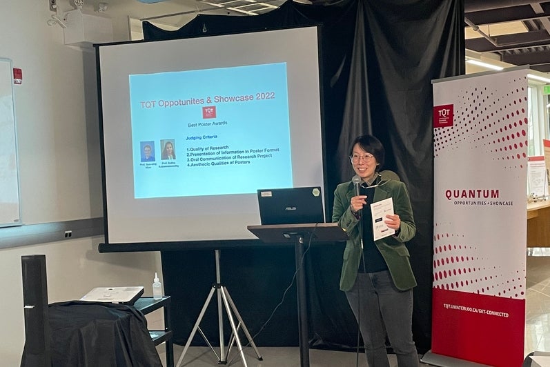 A person standing in front of a red and white banner and a projector screen, giving a talk. 