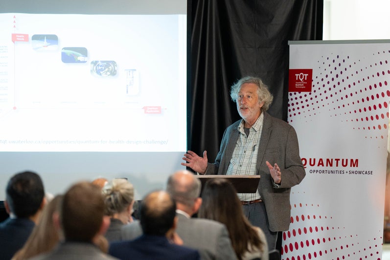 A person standing in front of a red and white banner and a projector screen, giving a talk. 
