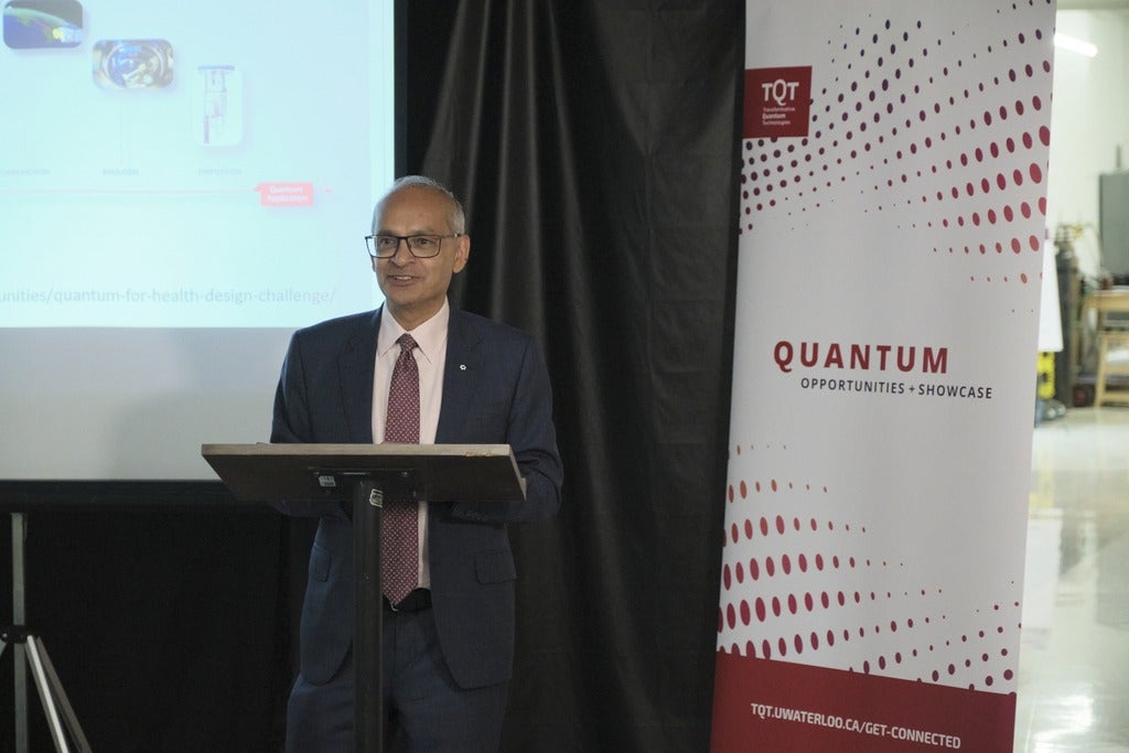 A person standing in front of a red and white banner and a projector screen, giving a talk. 