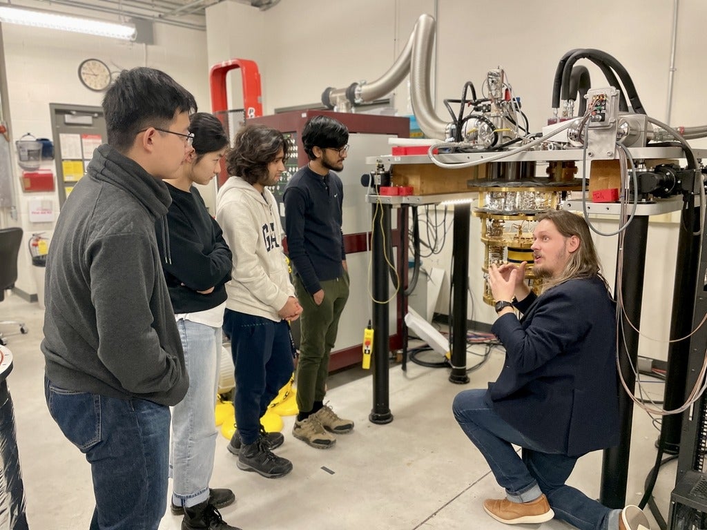 A person crouching on the ground, describing the Dilution Refrigerator to 4 standing people. 