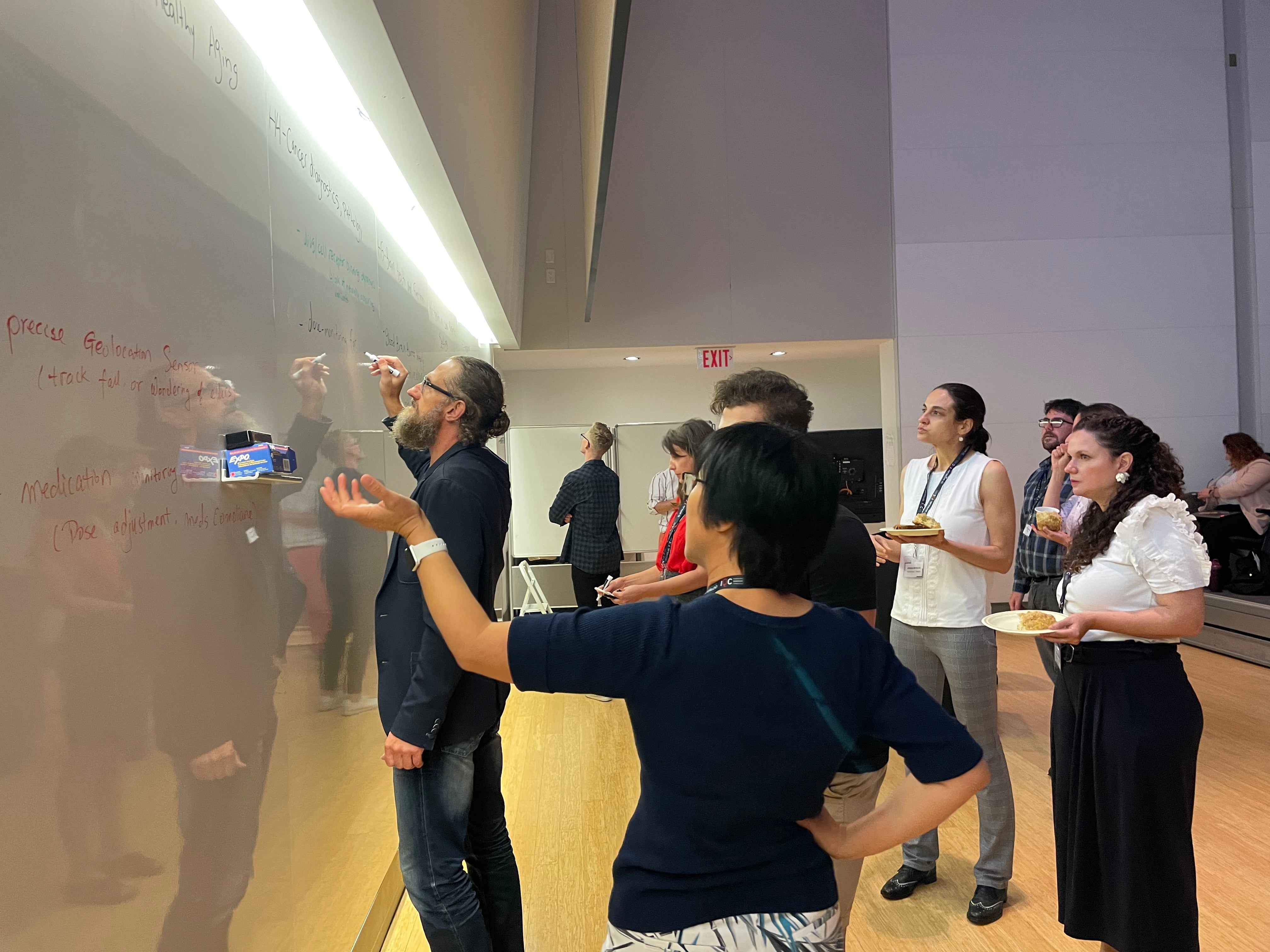 Group of people gathered around a whiteboard, while discussing and writing ideas down.