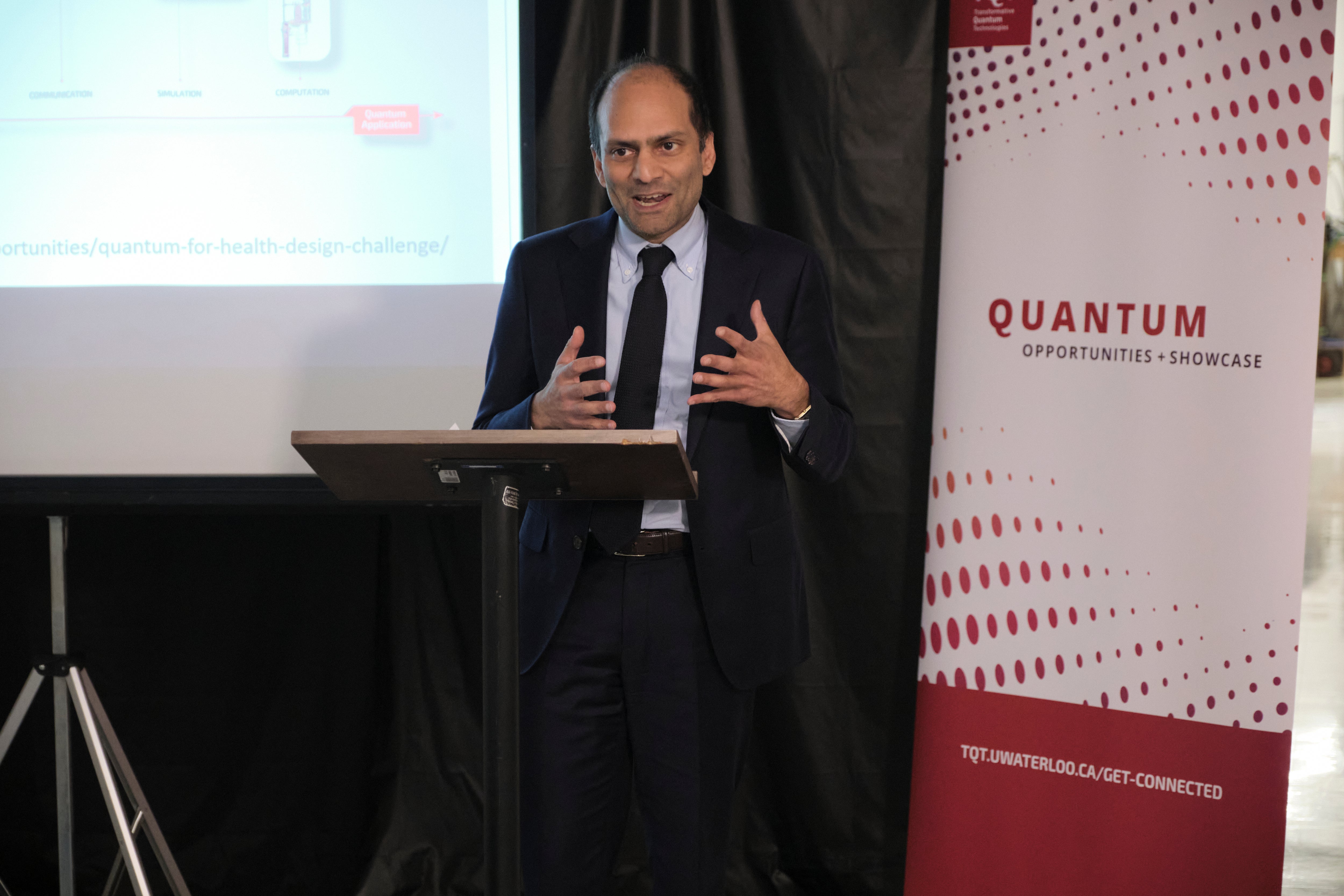 A person standing in front of a red and white banner and a projector screen, giving a talk. 