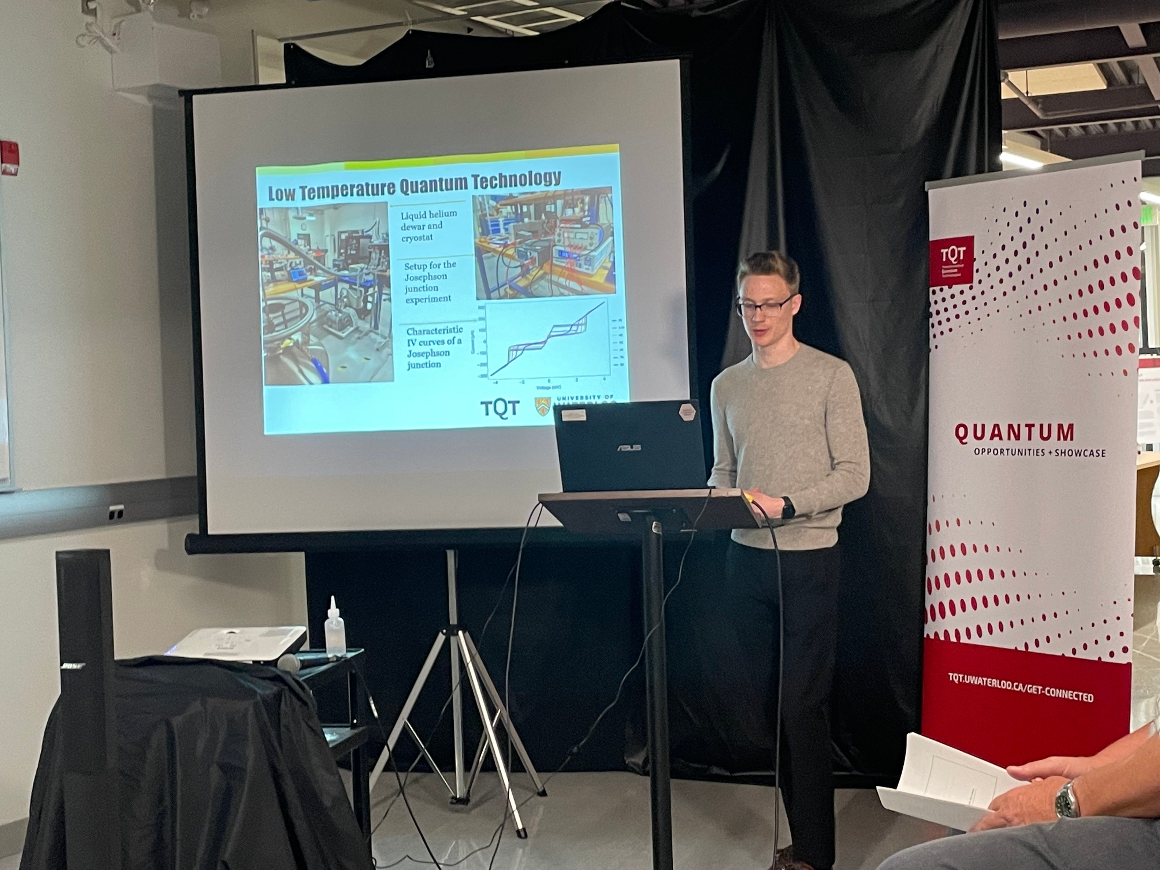 A person standing in front of a red and white banner and a projector screen, giving a talk. 