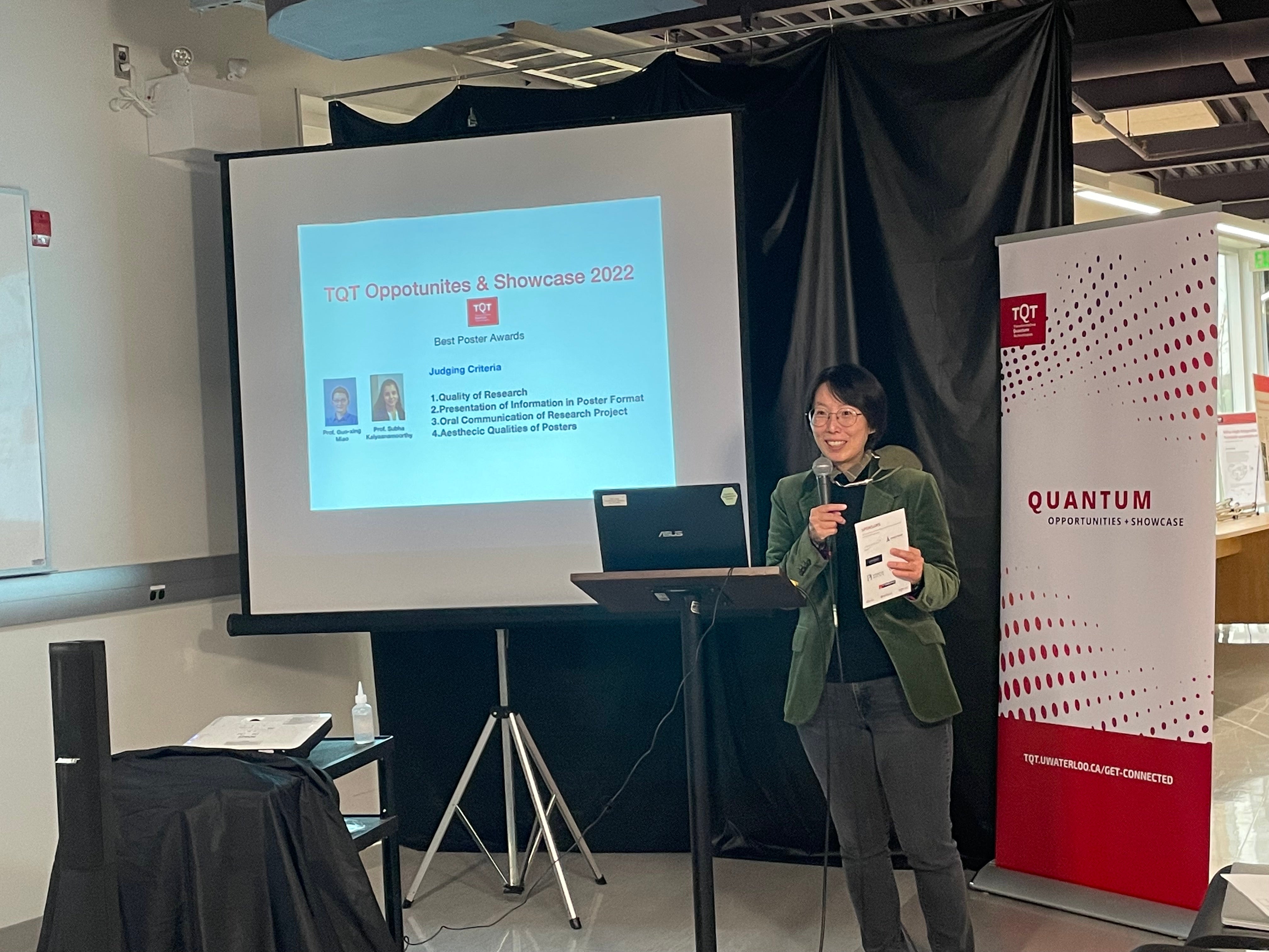 A person standing in front of a red and white banner and a projector screen, giving a talk. 