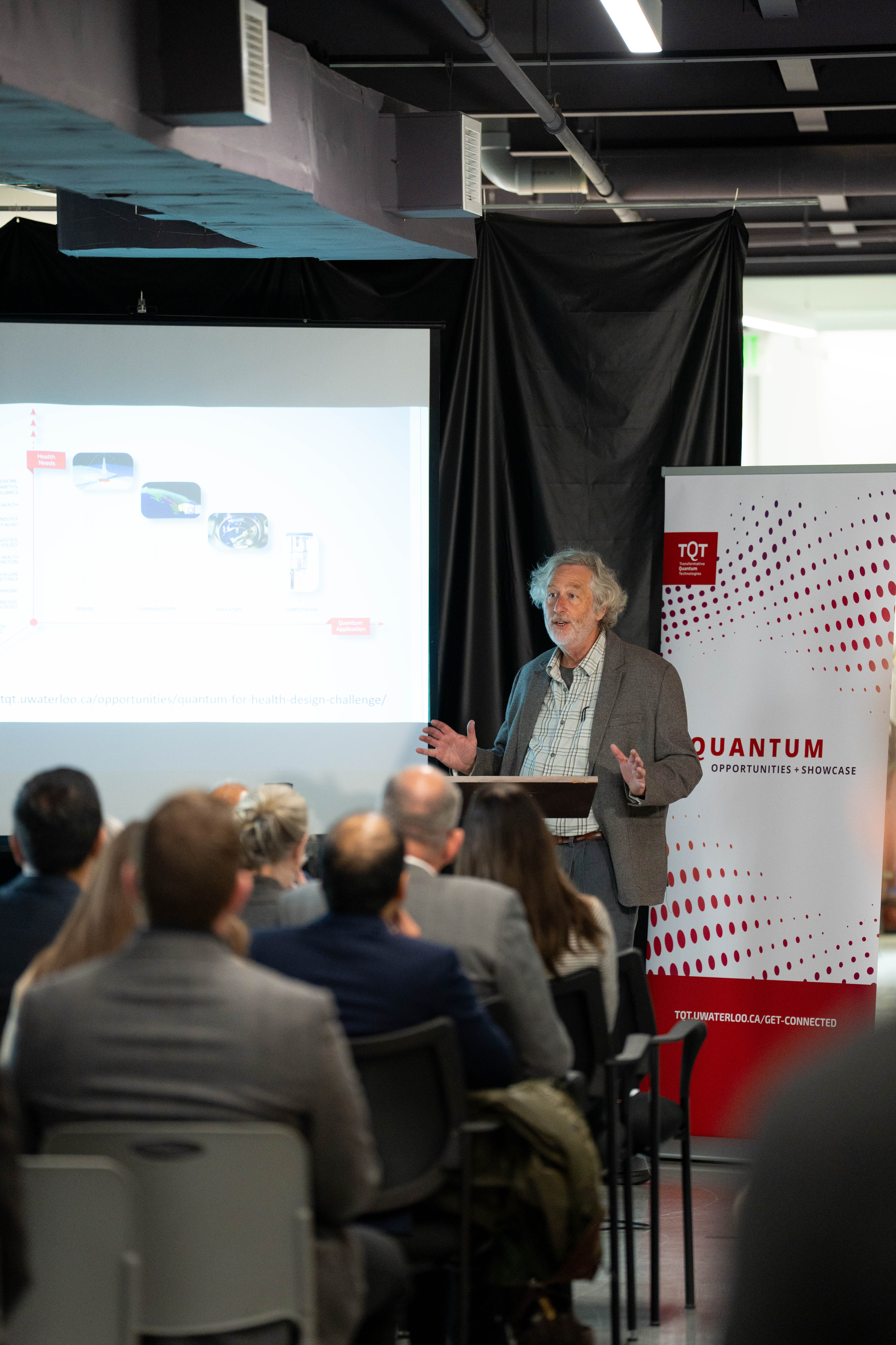 A person standing in front of a red and white banner and a projector screen, giving a talk. 