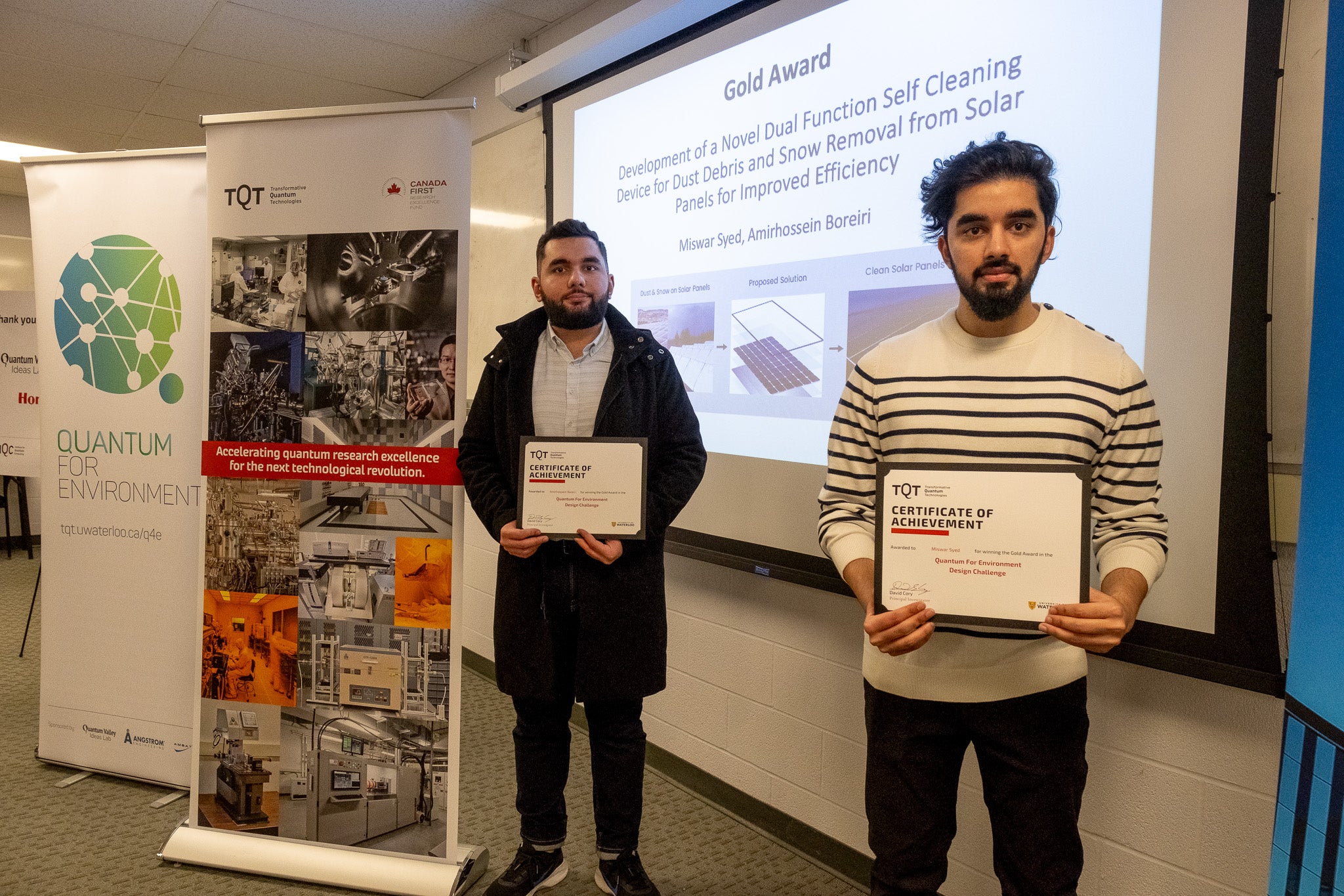 Two people standing in front of banners and a projector screen, while holding certificates
