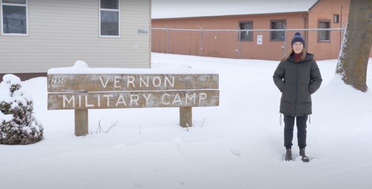 Megan Hamilton standing beside Vernon Military Camp sign in winter in front of two buildings