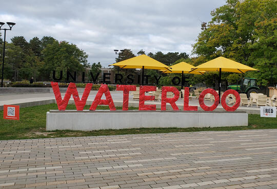The University of Waterloo sign, wrapped in red, with the Arts Quad int he background