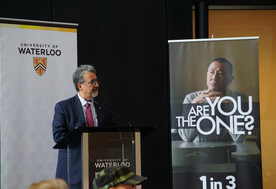 President Feridun Hamdullahpur presenting at the United Way launch next to a poster asking 