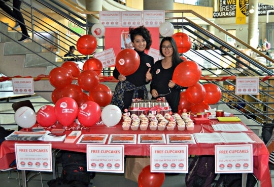 Two people stand in front of a red and white booth with balloons and cupcakes