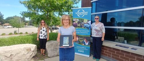 Three VAC volunteers stand outside, and one is holding a piece of artwork.