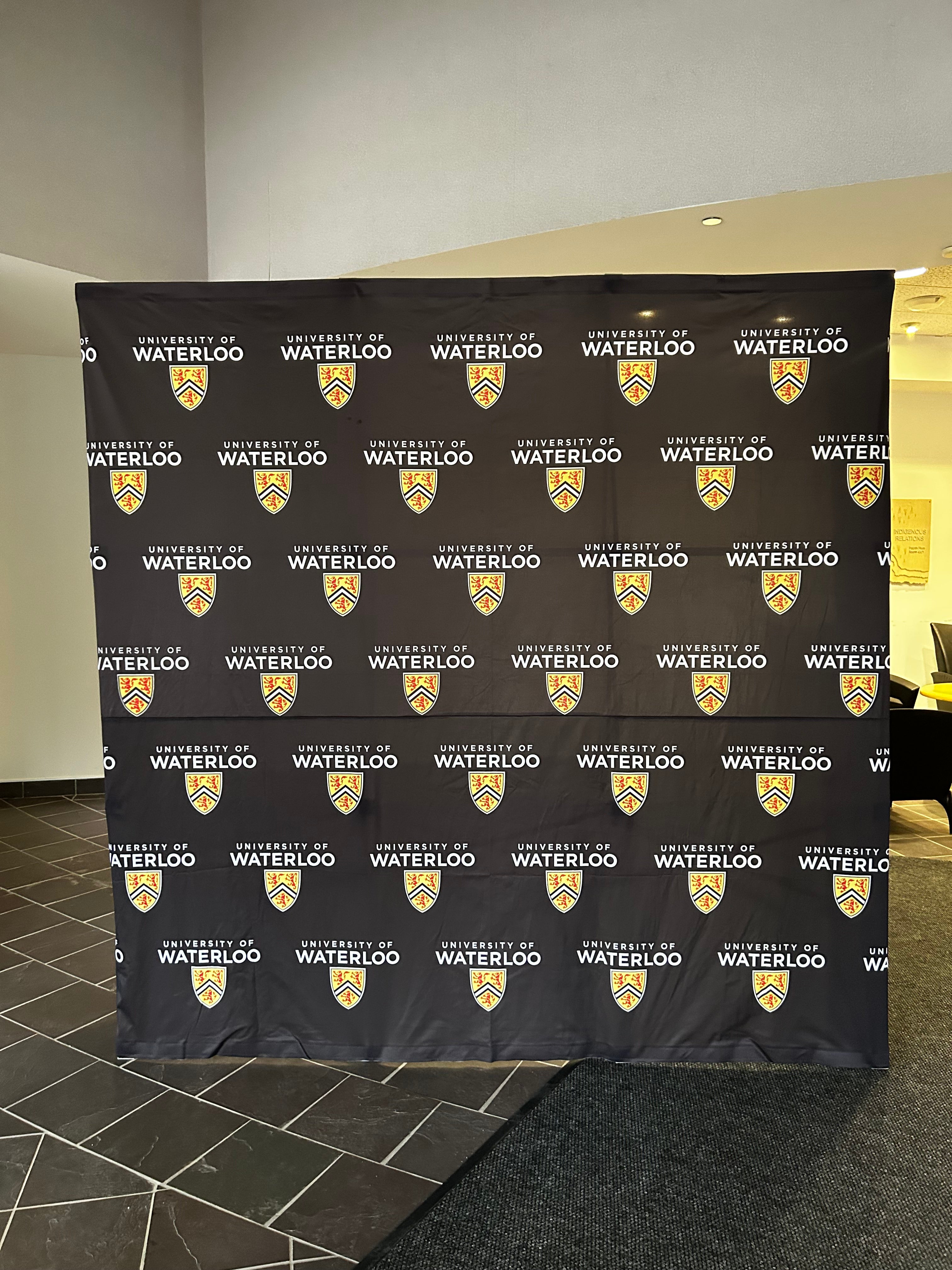 Black backdrop banner featuring a repeating pattern of the “University of Waterloo” name and crest logo.
