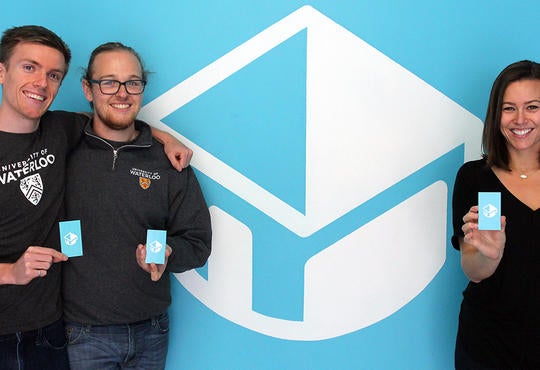 Jack Forbes, Zach Lima, and Courtney Sabo standing with business cards in front o company logo