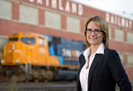 Corina Moore stands in front of stopped train on a railway and Northland Railway building